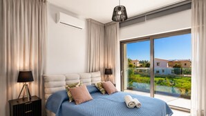 Een slaapkamer met een groot bed, een nachtkastje, een lamp, een airconditioner en uitzicht op een balkon met groen en gebouwen.