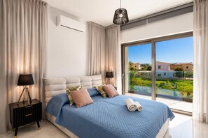 Ein Schlafzimmer mit einem großen Bett, einem Nachttisch, einer Lampe, einer Klimaanlage und Blick auf einen Balkon mit Grünpflanzen und Gebäuden.