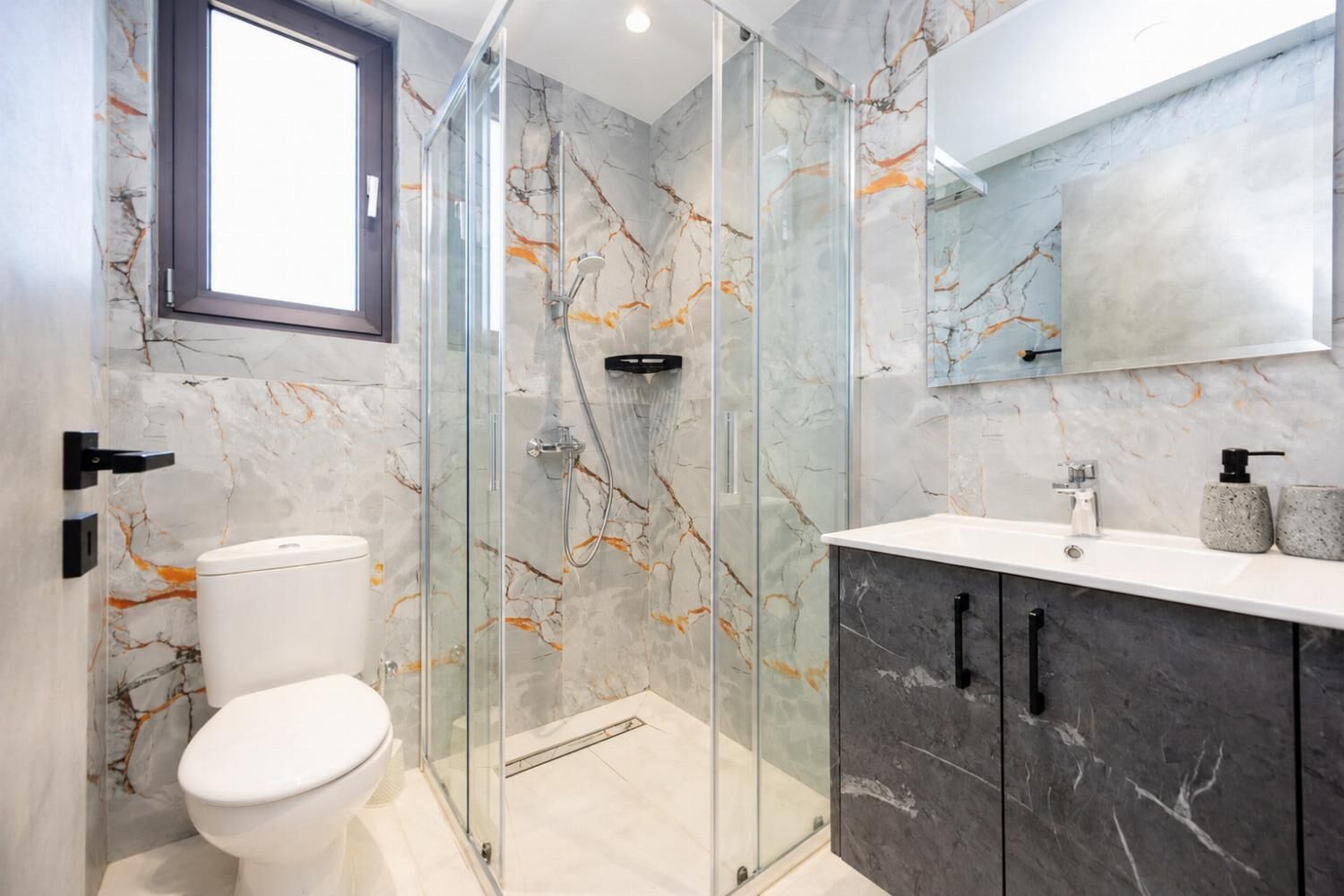 A modern bathroom with a glass shower enclosure, a toilet, a sink with a marble countertop, and a large mirror above the sink.