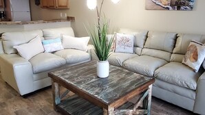 Sala de estar com mesa de centro de madeira, dois sofás, um vaso de planta e um quadro com uma cena de praia.