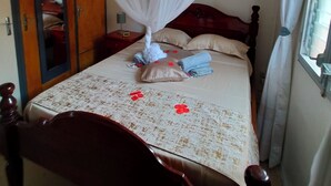 Quarto com cama, cabeceira de madeira, mesa de cabeceira, criado-mudo, espelho, guarda-roupa e janela com cortinas.