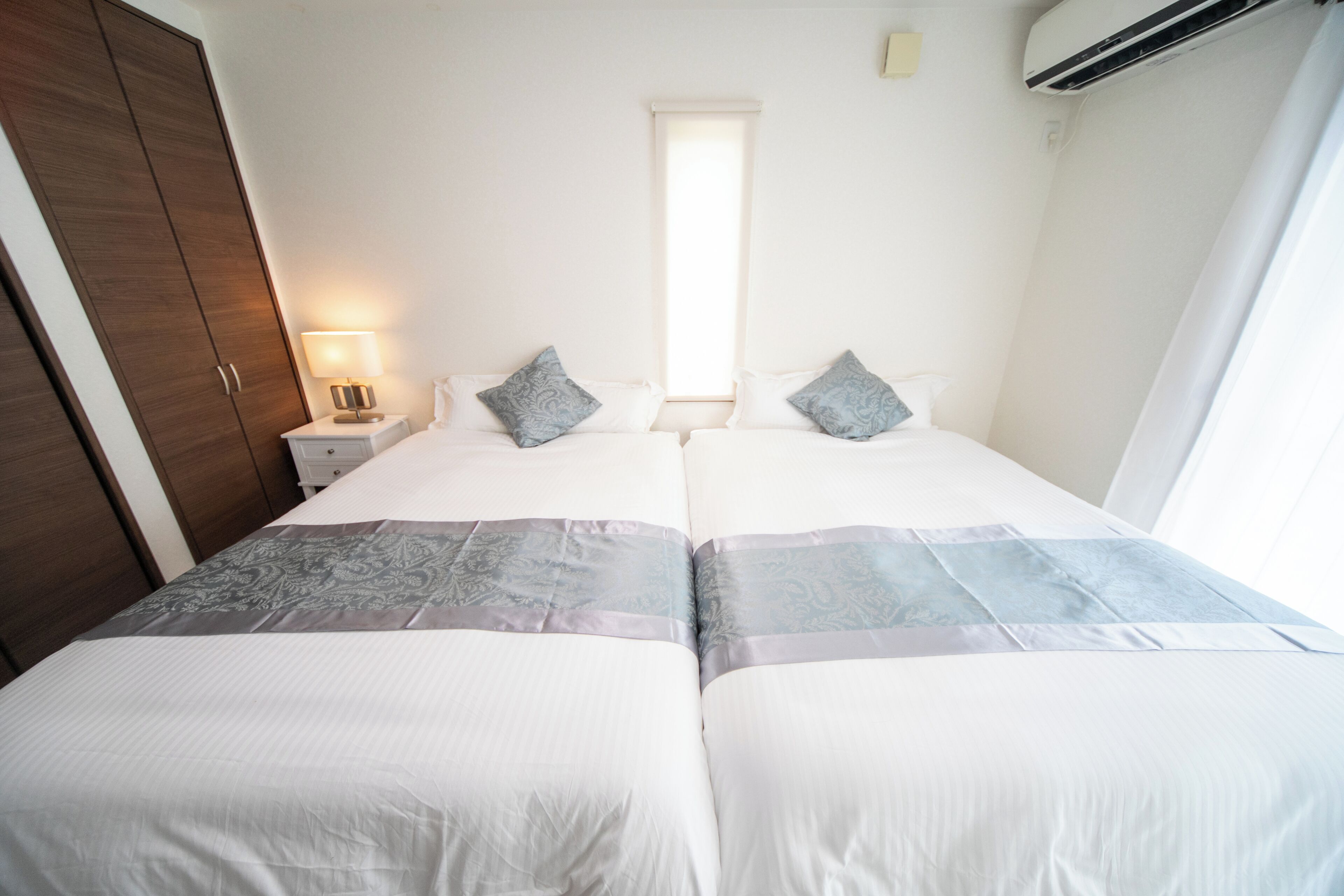 Een hotelkamer met twee bedden, een nachtkastje met lamp, een kledingkast en een raam met jaloezieën.