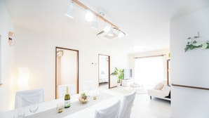A modern dining area with a long table, white chairs, and a chandelier. There is a sofa and a television in the background.