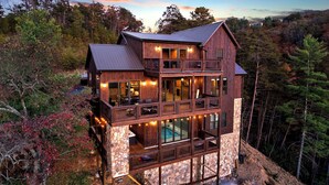 Une cabane en bois à plusieurs niveaux, dotée de fondations en pierre, de grandes fenêtres et d’un balcon donnant sur une zone boisée.