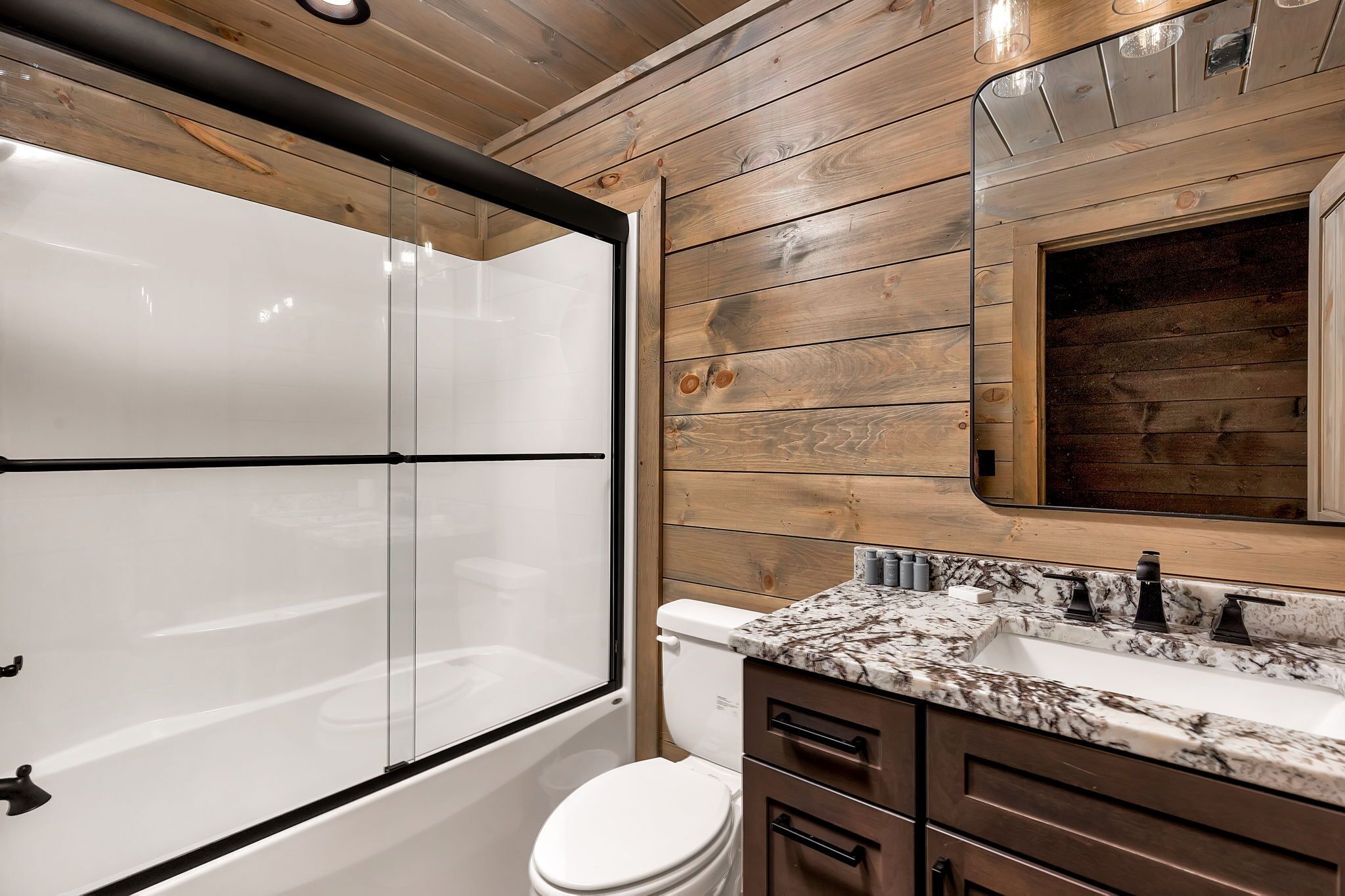 Un baño con ducha envuelta en vidrio, inodoro blanco y lavamanos de mármol con muebles de tonos oscuros.