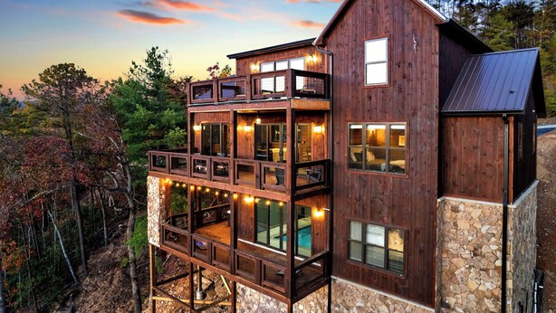 Une cabane en bois à plusieurs étages, dotée de fondations en pierre, de grandes fenêtres et d’un balcon donnant sur une zone boisée.