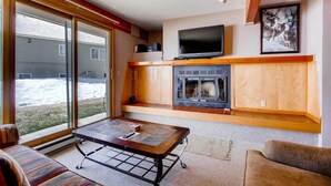 A living room with a fireplace, a television, and a large sliding glass door leading to a snowy yard. - Cozy Condo | Stunning Mountain Views + Fireplace (Crested Butte)