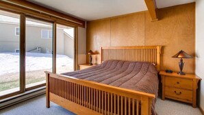 A bedroom with a wooden bed, two bedside tables with lamps, and a large window with a view of a house and yard. - Cozy Condo | Stunning Mountain Views + Fireplace (Crested Butte)