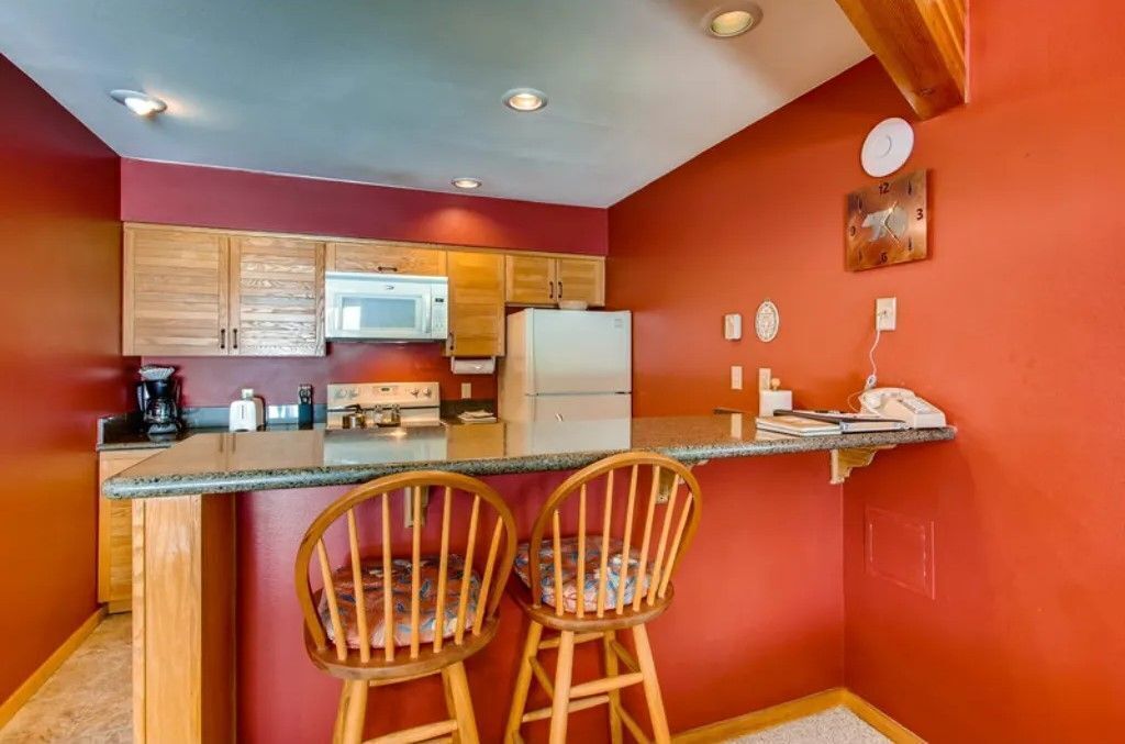 A compact kitchen with wooden cabinets, a microwave, a refrigerator, and a small bar area with two chairs.