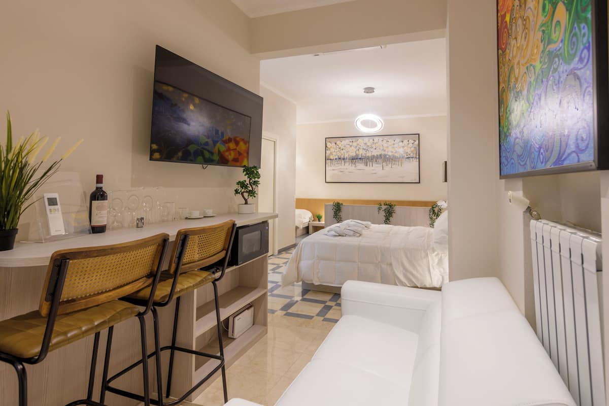 A modern hotel room with a kitchenette, a dining area with bar stools, a bed, and a large window.