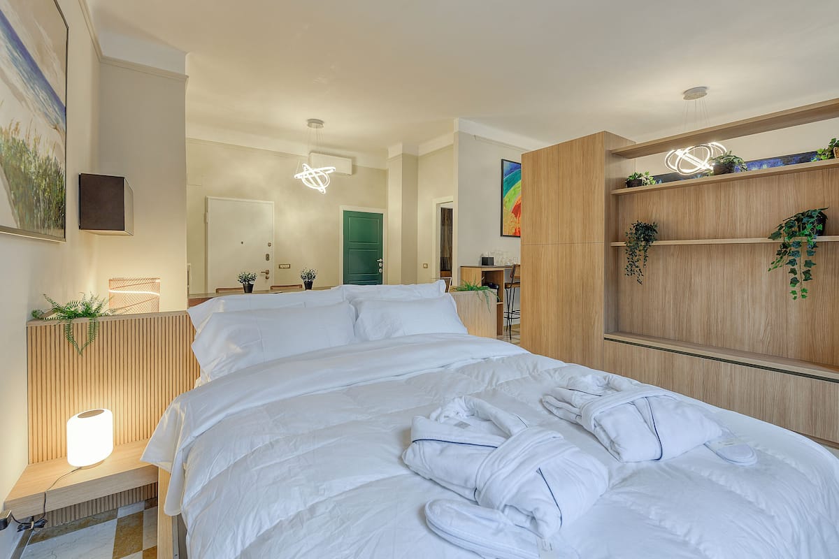 A modern hotel room with a large bed, wooden headboard, a bedside table with a lamp, a mirror, and a wall-mounted TV.