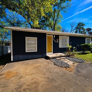 A single-story house with a dark exterior, a yellow door, and a small front yard with a gravel path and a few plants.