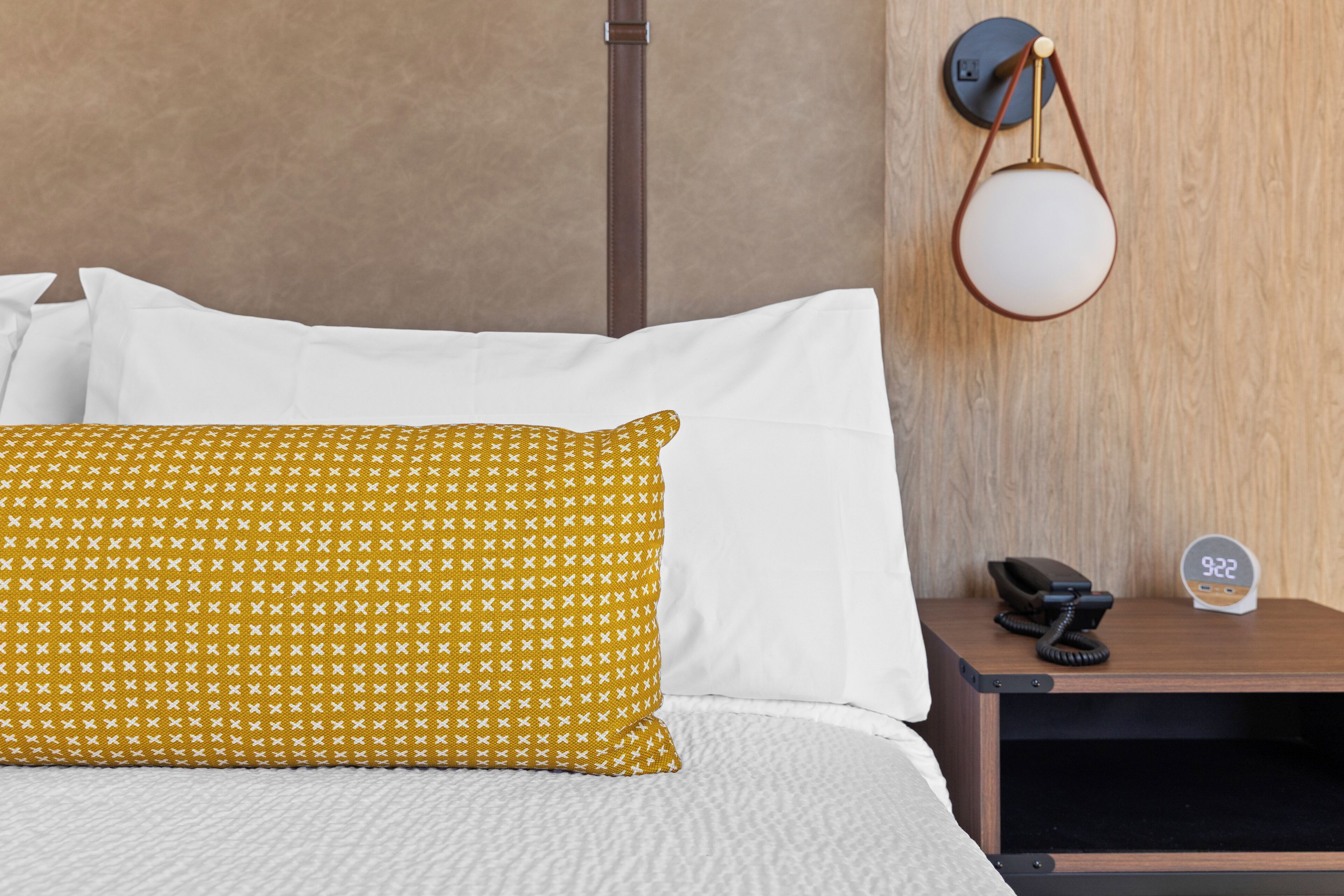 A bed with white and yellow patterned pillows, a bedside table with a phone and a clock, and a wall-mounted lamp.
