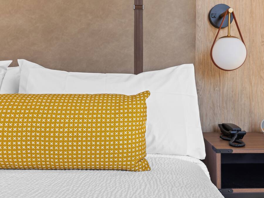 A bed with white and yellow patterned pillows, a bedside table with a phone and a clock, and a wall-mounted lamp.