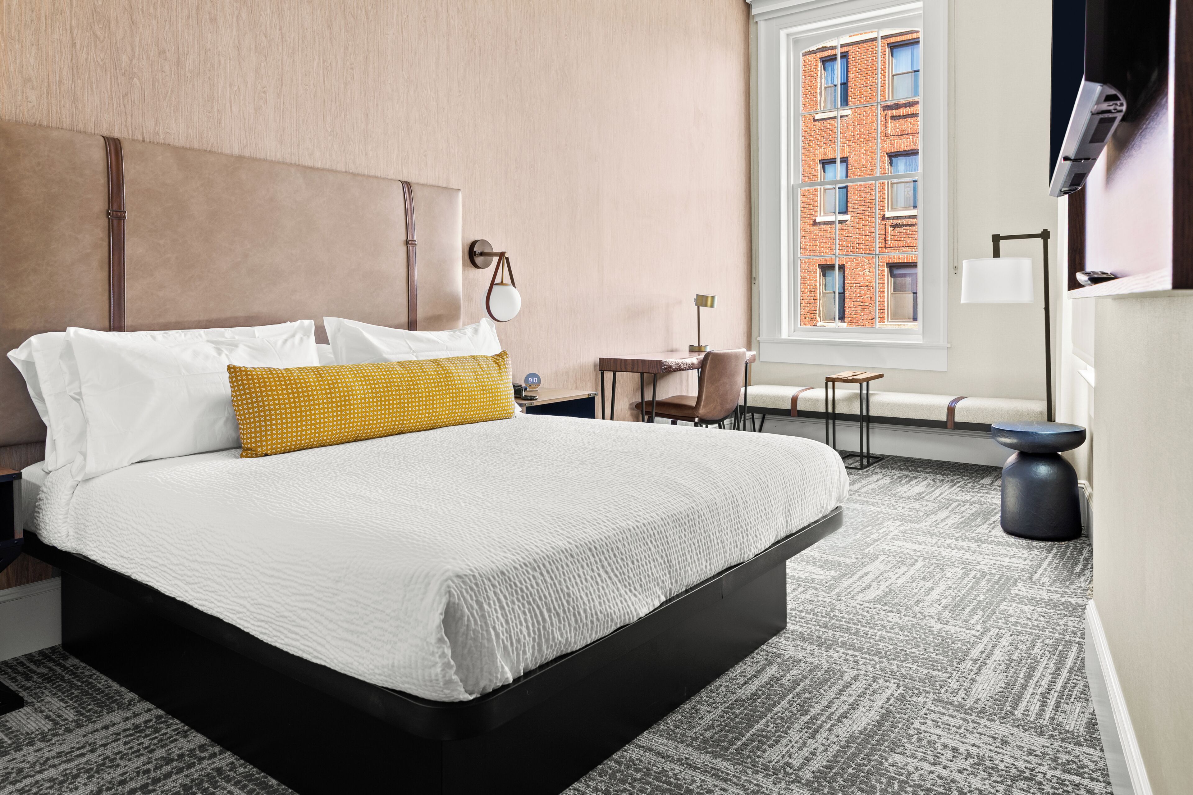 Ein Hotelzimmer mit einem großen Bett, einem kleinen Tisch, einem Sessel und Blick auf ein Backsteingebäude durch das Fenster.