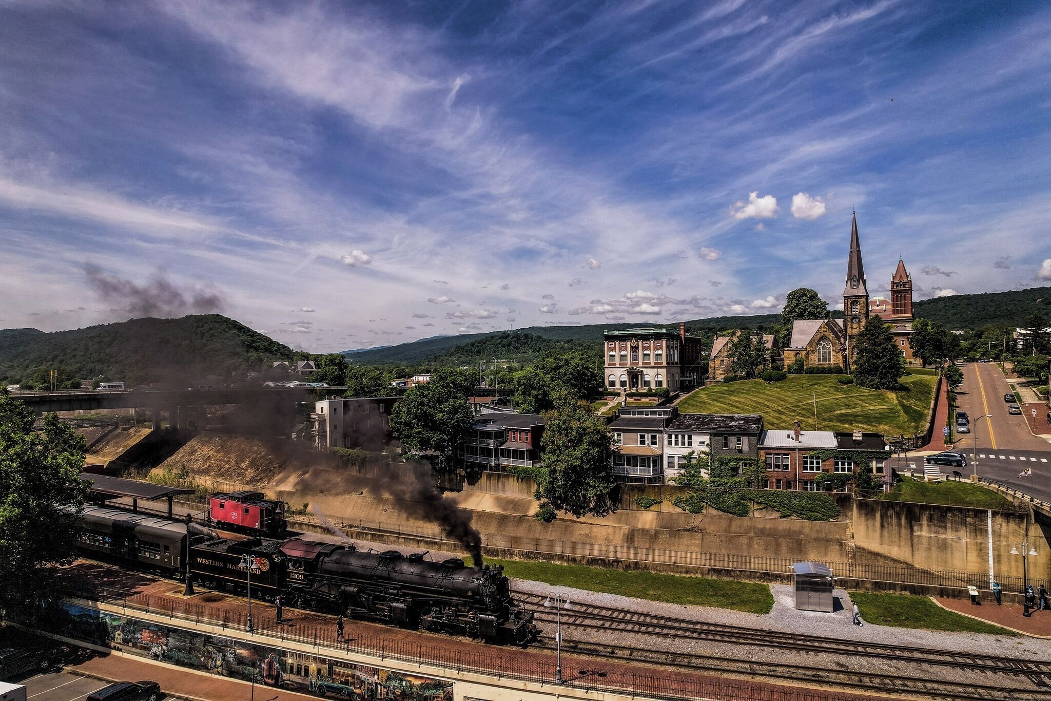 A train station with a steam locomotive, a church with a tall spire, and a bridge in the background.