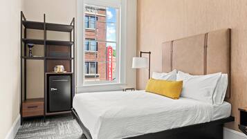 A hotel room with a bed, a desk, a lamp, a wardrobe, and a window with a view of a brick building.