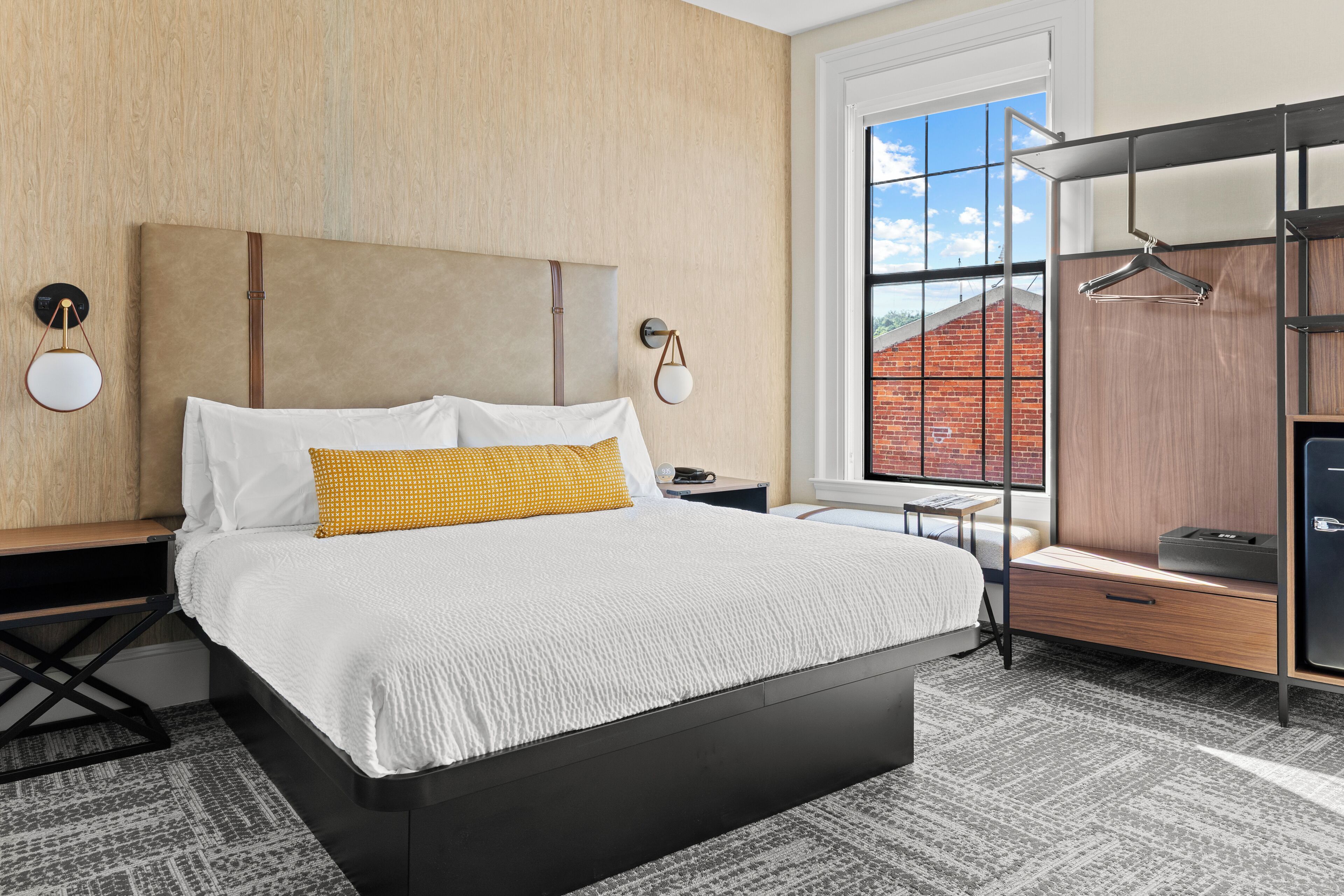 A modern hotel room with a large bed, a desk, a wardrobe, and a window with a view of a brick building.