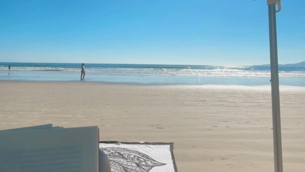 En strandscene med en person, der ligger under en parasol, et håndklæde med bladmotiv og en strandstol.