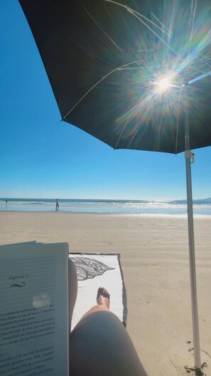 Eine Strandansicht mit einer Person, die unter einem Strandsonnenschirm liegt, einem Handtuch mit Blattmuster und einem Liegestuhl.