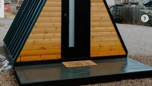 Uma pequena cabana de madeira triangular com porta preta e janela, com montanhas ao fundo.