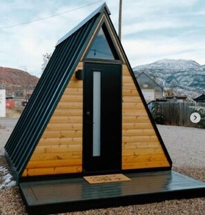 Una pequeña cabaña de madera triangular con una puerta negra y una ventana, con montañas al fondo.