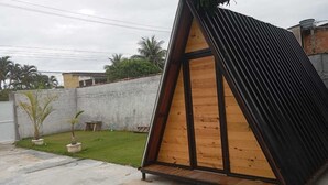 Uma estrutura triangular com interior de madeira e exterior preto, localizada em um gramado com um muro de concreto e uma pequena mesa ao fundo.