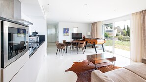 Uma cozinha moderna com forno embutido, sala de jantar com mesa redonda e cadeiras, uma janela grande com cortinas e um sofá de couro na sala de estar.