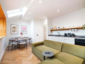 Ein moderner Wohnraum mit Essbereich, einem grünen Sofa, einem kleinen runden Tisch mit Fernbedienung und einer Küche mit Holzregal.