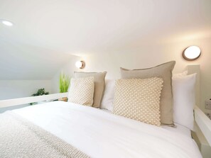 Een bed met witte beddengoed, twee gestructureerde kussens, een nachtkastje met een lamp en uitzicht op de zee.
