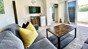 Un salon moderne comprenant un canapé, une table basse, un téléviseur à écran plat, un petit coin cuisine et une porte-fenêtre coulissante donnant sur un espace extérieur.