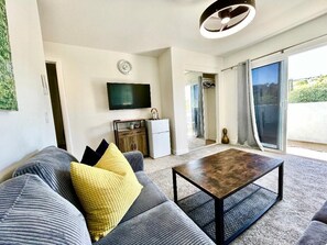 A modern living room with a sofa, a coffee table, a flat-screen TV, a small kitchen area, and a sliding glass door leading to an outdoor area. -  
Charming Cozy Suite in Poway with Sloped Ceiling & Private Balcony (Poway)
