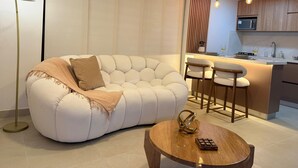 Una sala de estar moderna con un sofá blanco, una mesa de centro redonda y un comedor con sillas de madera, con una cocina al fondo.