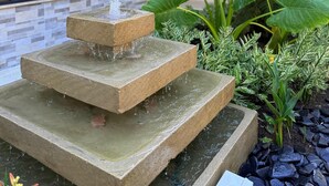 Una fuente de piedra escalonada con agua en cascada, rodeada de exuberantes plantas verdes y un edificio moderno con fachada de ladrillo y grandes ventanales.