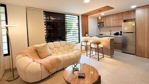 Una sala de estar moderna con un sofá y una mesa de centro, una cocina con armarios de madera y electrodomésticos de acero inoxidable, y un ventanal con persianas.