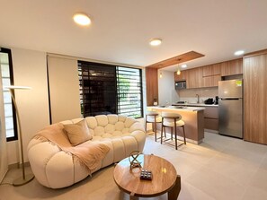 A modern living room with a sofa and a coffee table, a kitchen with wooden cabinets and stainless steel appliances, and a large window with blinds. - Discover modern and stylish apt in Limonar (Cali)