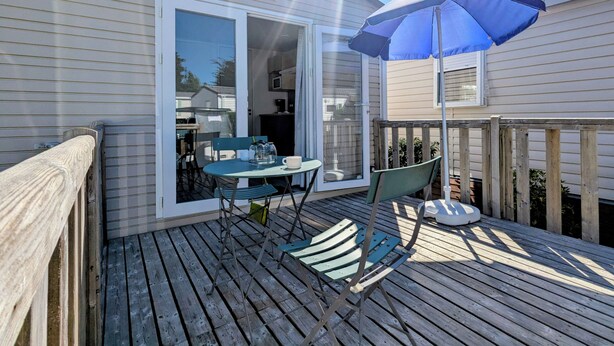 Une terrasse en bois avec une table et des chaises, un parasol bleu et une porte-fenêtre coulissante donnant sur un coin cuisine.