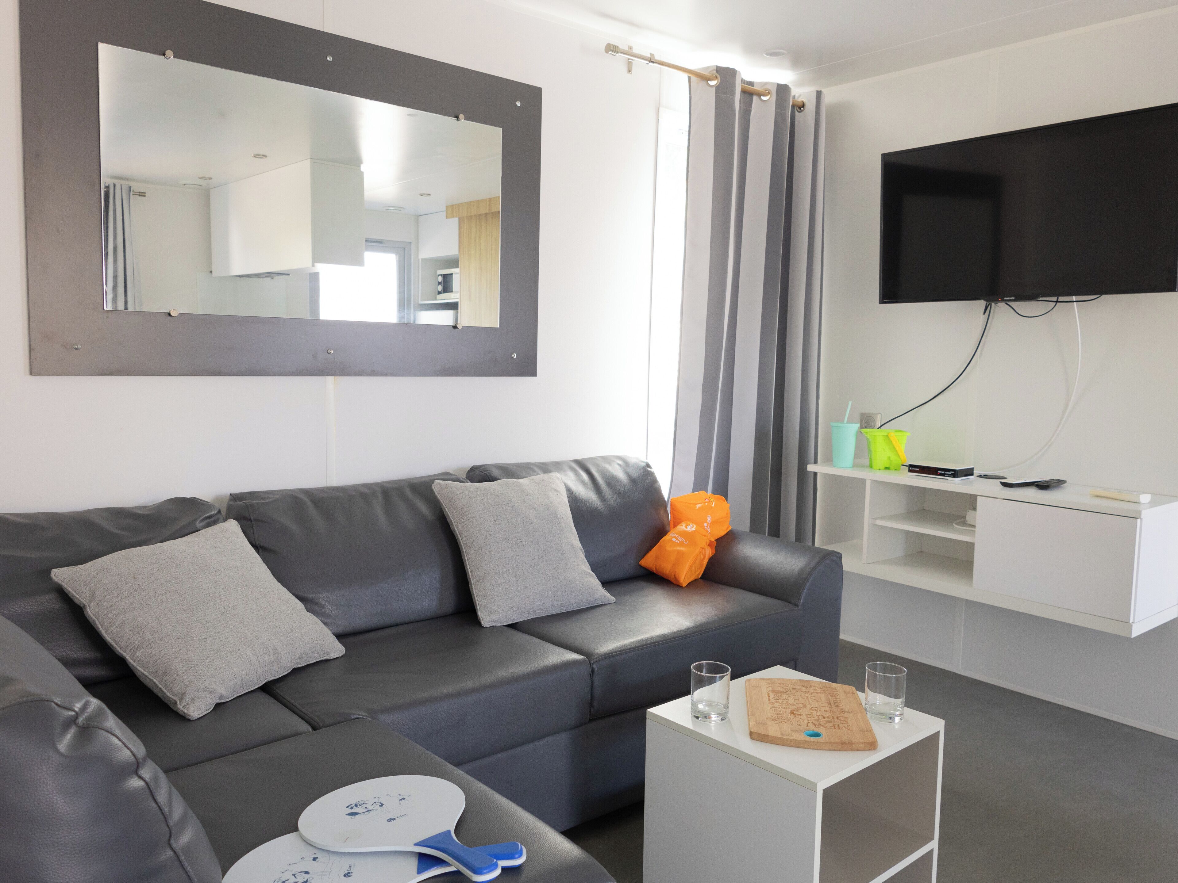 A modern living room with a grey leather sofa, a flat-screen TV mounted on the wall, and a small white side table with a cutting board and glasses.