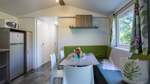 Et kompakt køkken med mikrobølgeovn, køleskab og et spiseområde med bord og stole. Der er en grøn sofa med pude og et vindue med gardiner.
