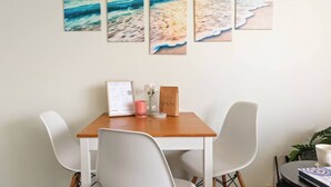 A dining area with a wooden table, white chairs, and a wall-mounted artwork depicting a beach scene. - Shoreleaf Haven Moments from Waves and Seaside Eats (Dee Why)