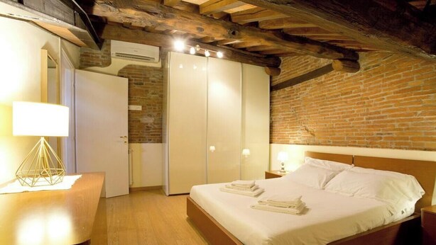 A bedroom with a wooden bed, a nightstand, a lamp, a wardrobe, and a brick wall. - Casa San Girolamo, historic flat with terrace in Lucca's City Center (Lucca)
