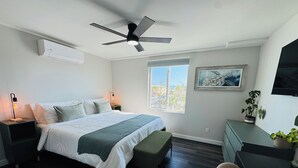 Un dormitorio con cama, mesitas de noche, escritorio, silla, ventilador de techo, ventana y un cuadro en la pared.