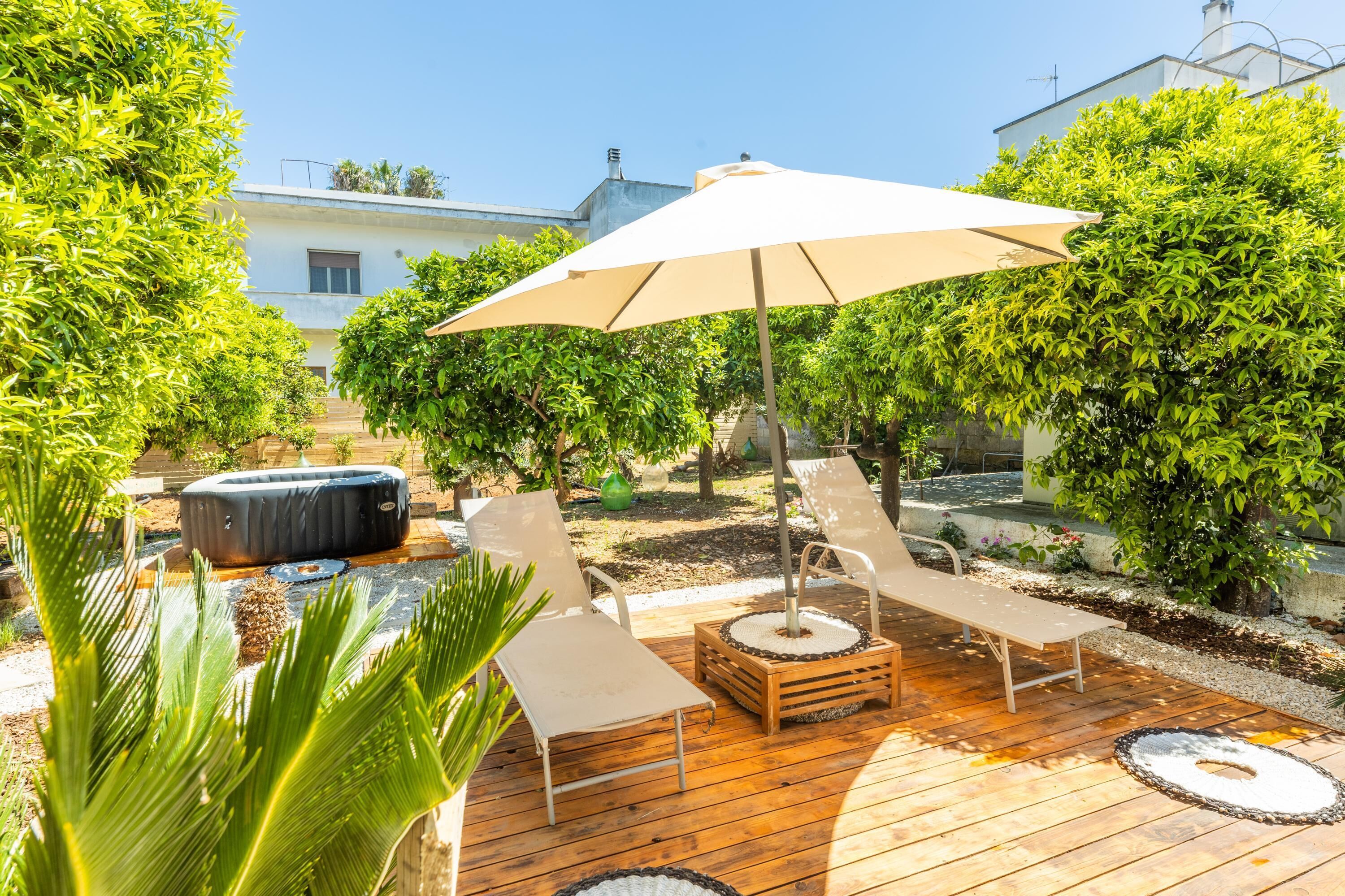 A wooden deck with lounge chairs and a large umbrella, surrounded by greenery and a hot tub.