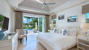 A spacious bedroom with a large bed, a desk with a chair, a TV, and a view of the outdoors. - 4 bed Prestigious Luxury Villa, Royal Westmoreland (Lancaster)