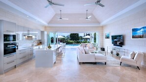 A spacious modern kitchen with a central island, dining area, and seating by a large window overlooking a pool. - 4 bed Prestigious Luxury Villa, Royal Westmoreland (Lancaster)