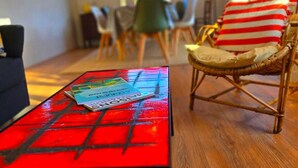 Un salon avec une table rouge, un fauteuil en osier avec des coussins à rayures, et un coin repas à l’arrière-plan.