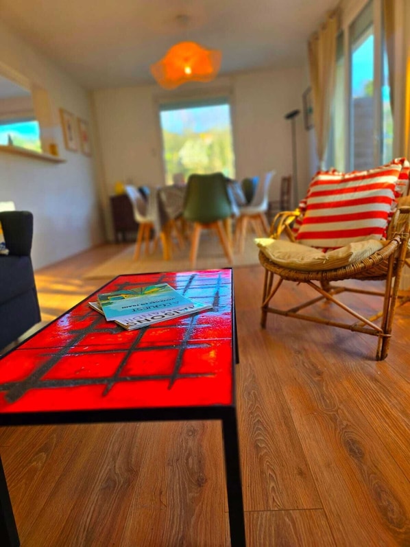 A living room with a red table, a wicker chair with striped cushions, and a dining area in the background.