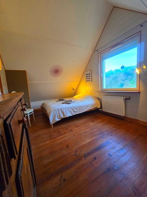 A bedroom with a sloped ceiling, a bed, a small table, a chair, and a window with a view of the outdoors.