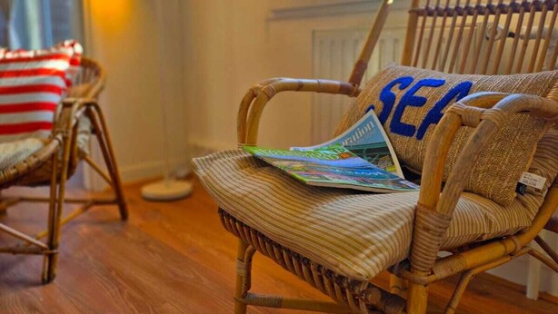 Une chaise en osier, un magazine et un coussin orné du mot « MER », un plancher en bois et une fenêtre donnant sur un paysage agréable.