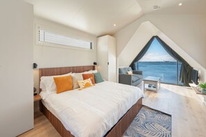 Ein modernes Schlafzimmer mit einem großen Bett, einer kleinen Sitzecke und Blick auf das Wasser.